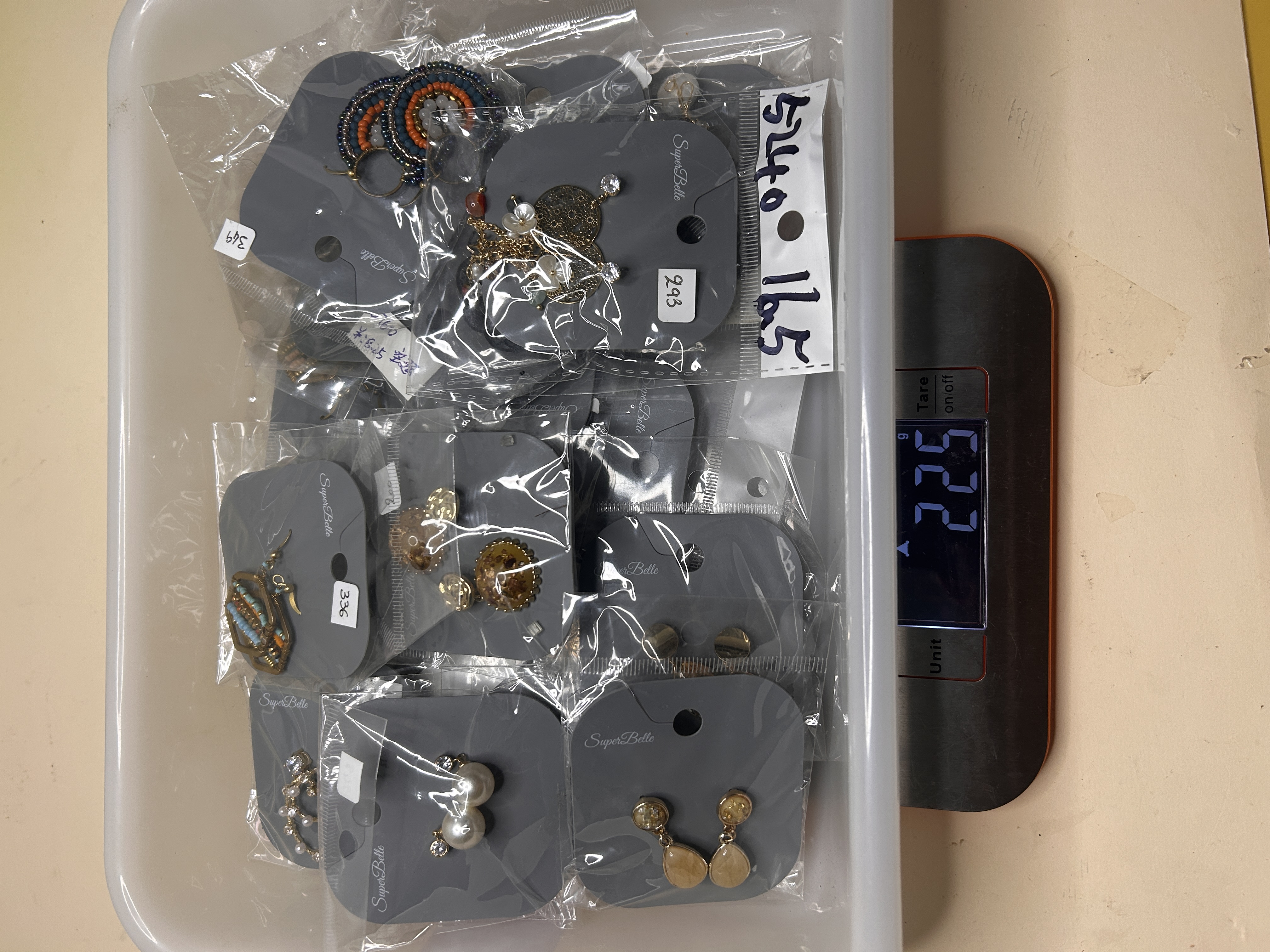 Vista cenital de una caja rectangular llena de varios pendientes de bisutería empaquetados individualmente, sobre una báscula que muestra 225g.