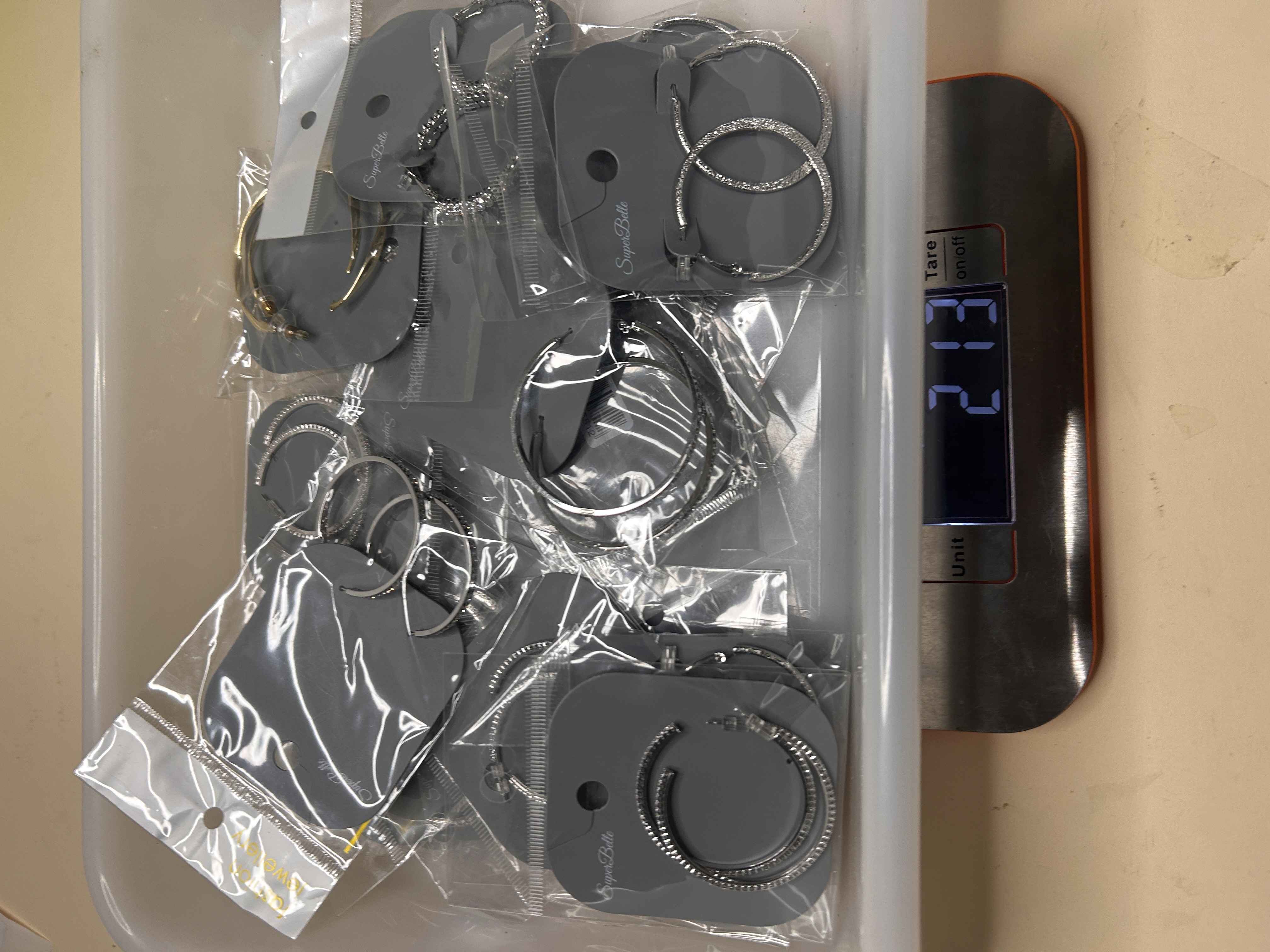 Vista cenital del lote al por mayor de pendientes de aro BO-101 en una bandeja blanca sobre una báscula digital que muestra 213 gramos, con varios estilos y acabados visibles en envases de plástico transparente.
