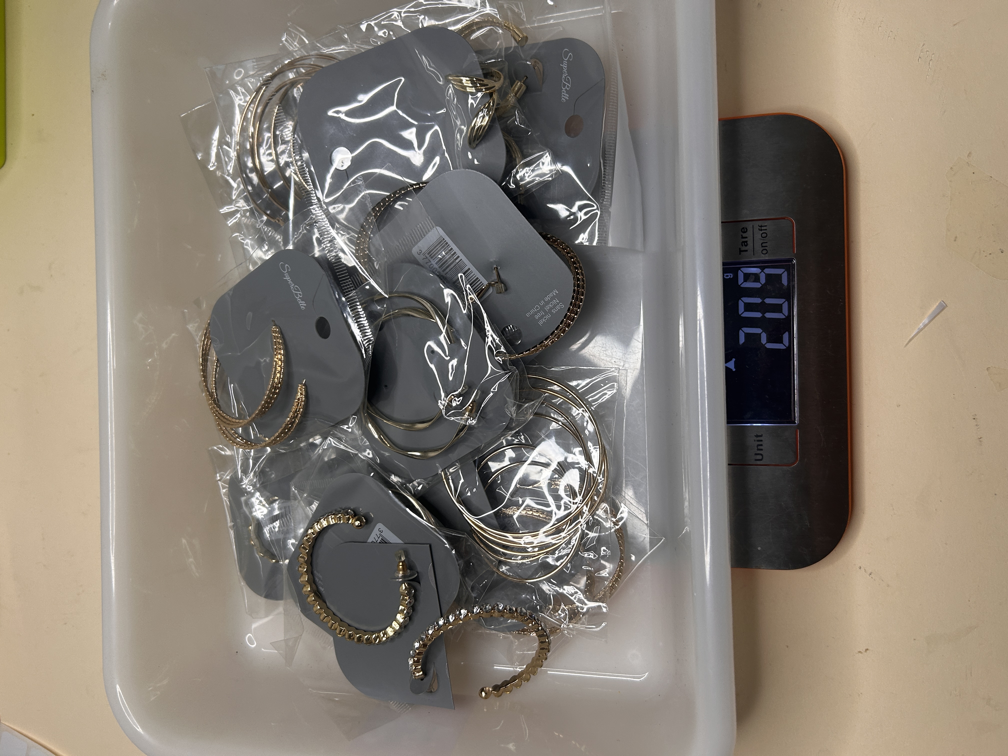 Vista superior de un contenedor lleno de varios pendientes de aro dorados, empaquetados individualmente en tarjetas grises, apoyado sobre una báscula digital que muestra '209g'.