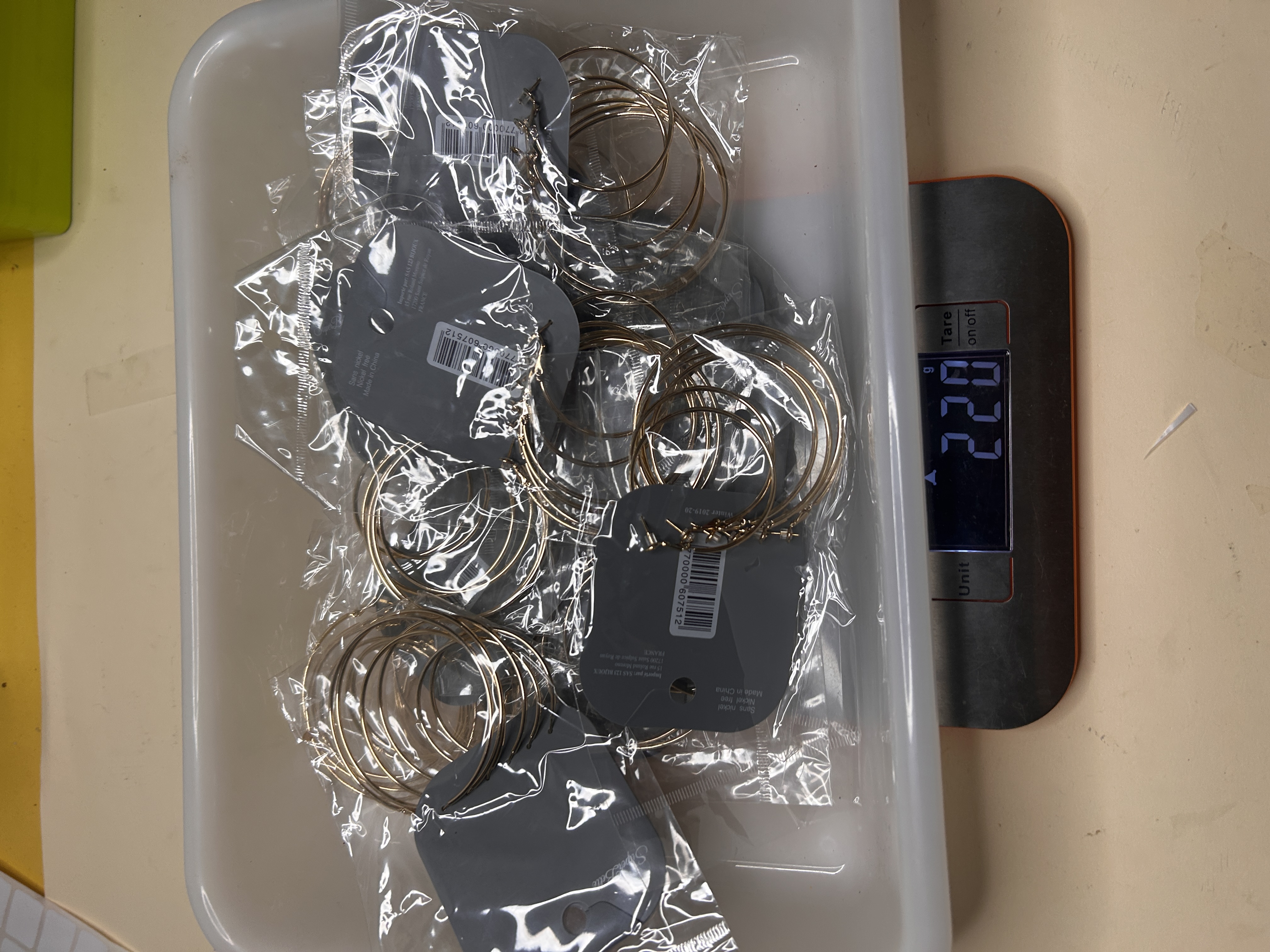 Vista superior de una bandeja llena de sets de pendientes de aro dorados empaquetados individualmente sobre una báscula que muestra 220 gramos.