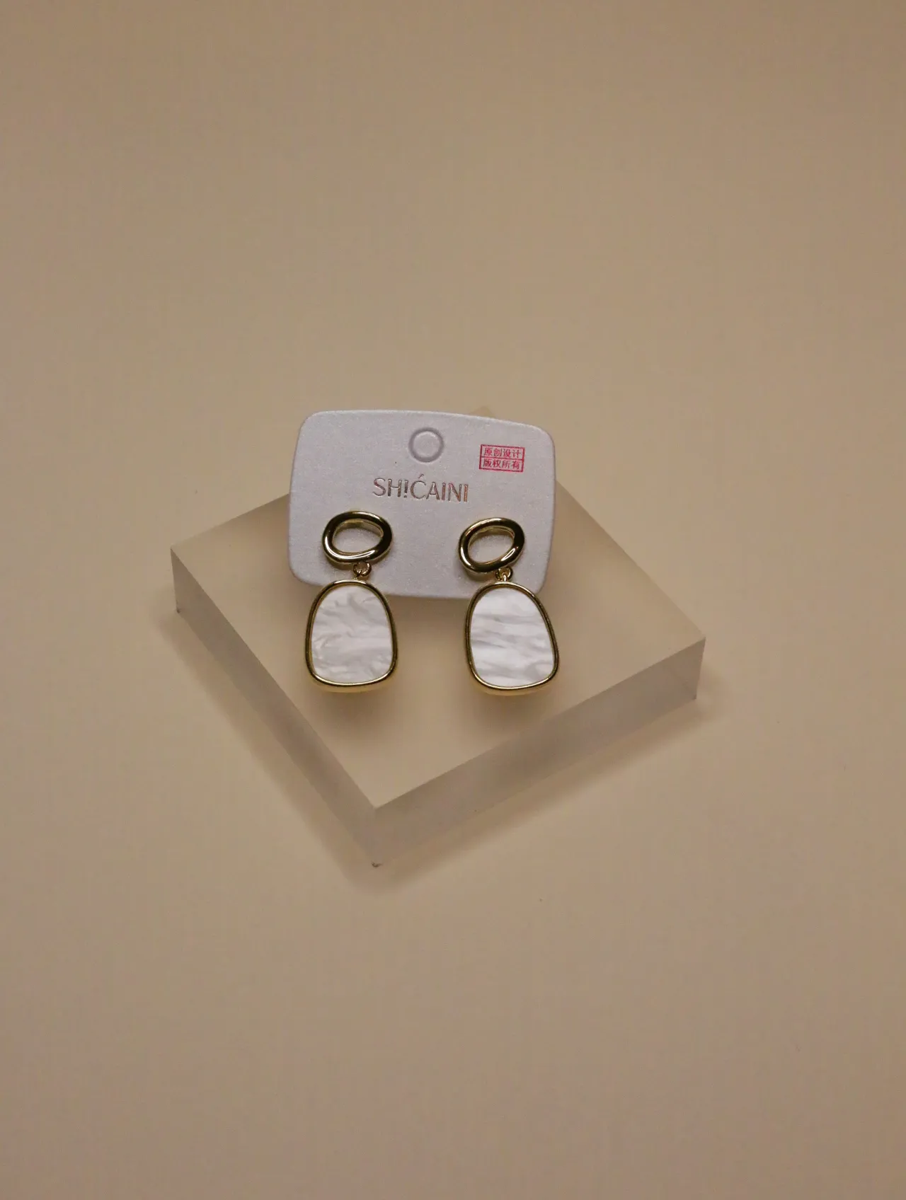 Un par de pendientes geométricos blancos con borde dorado expuestos en una tarjeta blanca y bloque transparente.