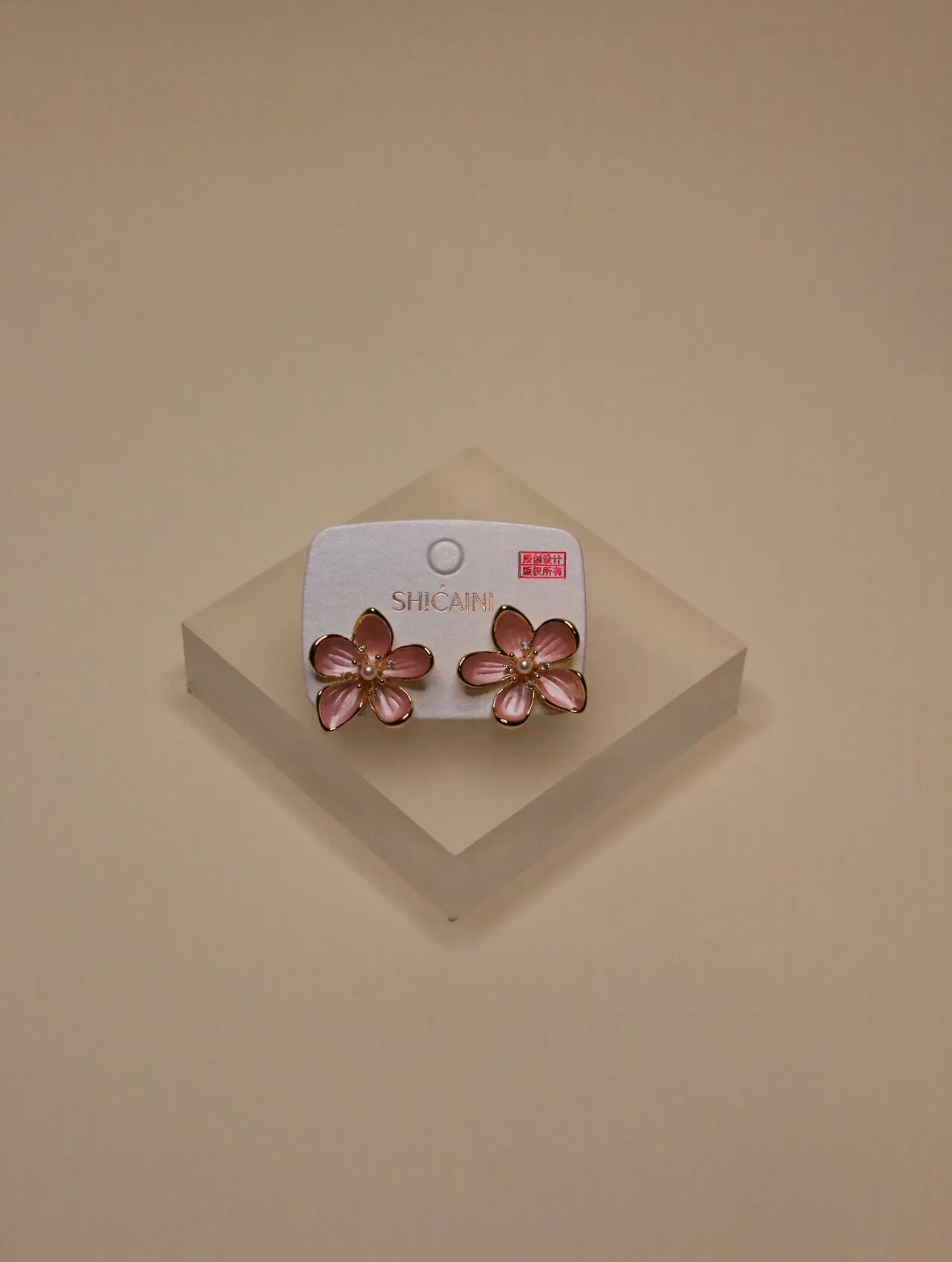 Primer plano de un par de pendientes de flor con esmalte rosa en una tarjeta de presentación blanca, sobre un bloque neutro.