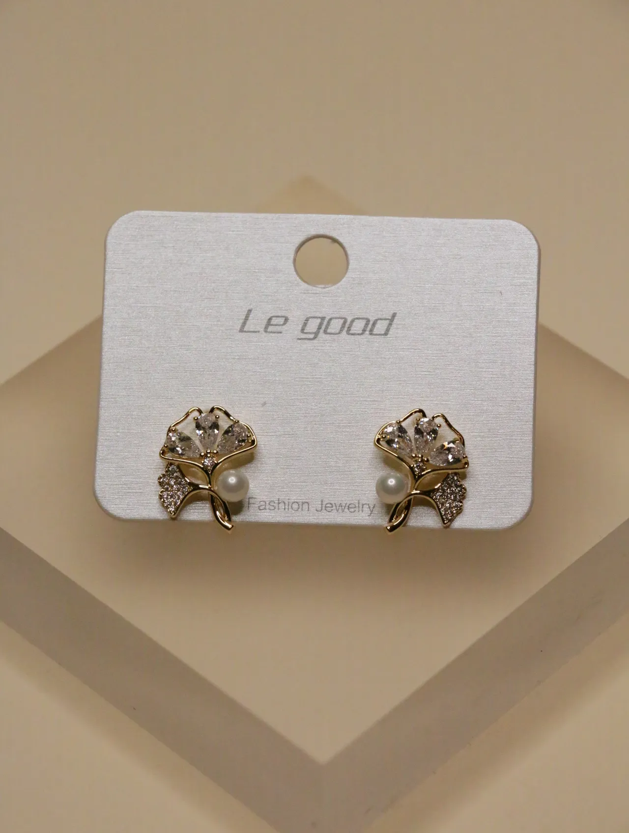 Un par de pendientes dorados con flores, perlas y circonitas expuestos en una tarjeta blanca.