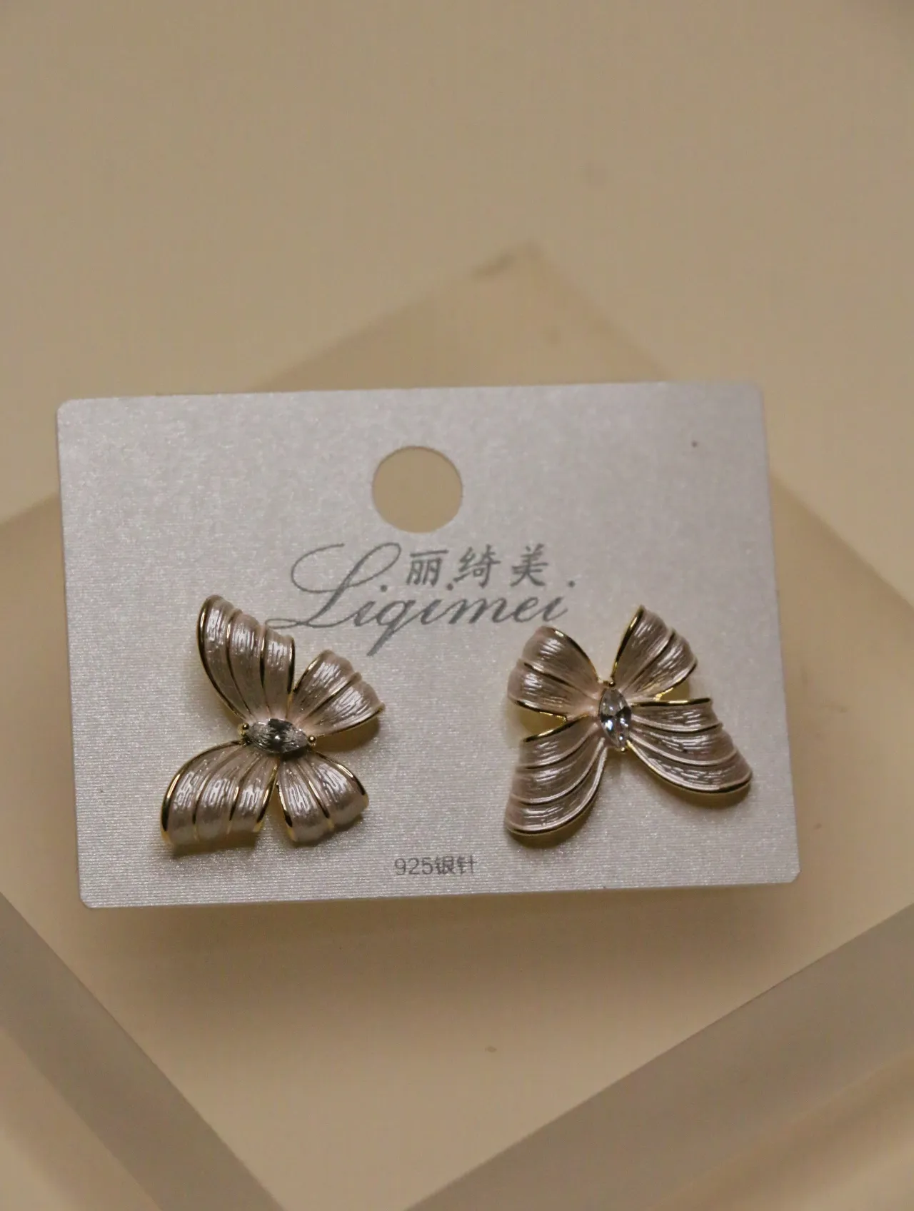 Un par de elegantes pendientes de lazo dorados y blancos en una tarjeta 'Liqimei', mostrando los detalles intrincados y el brillo central.