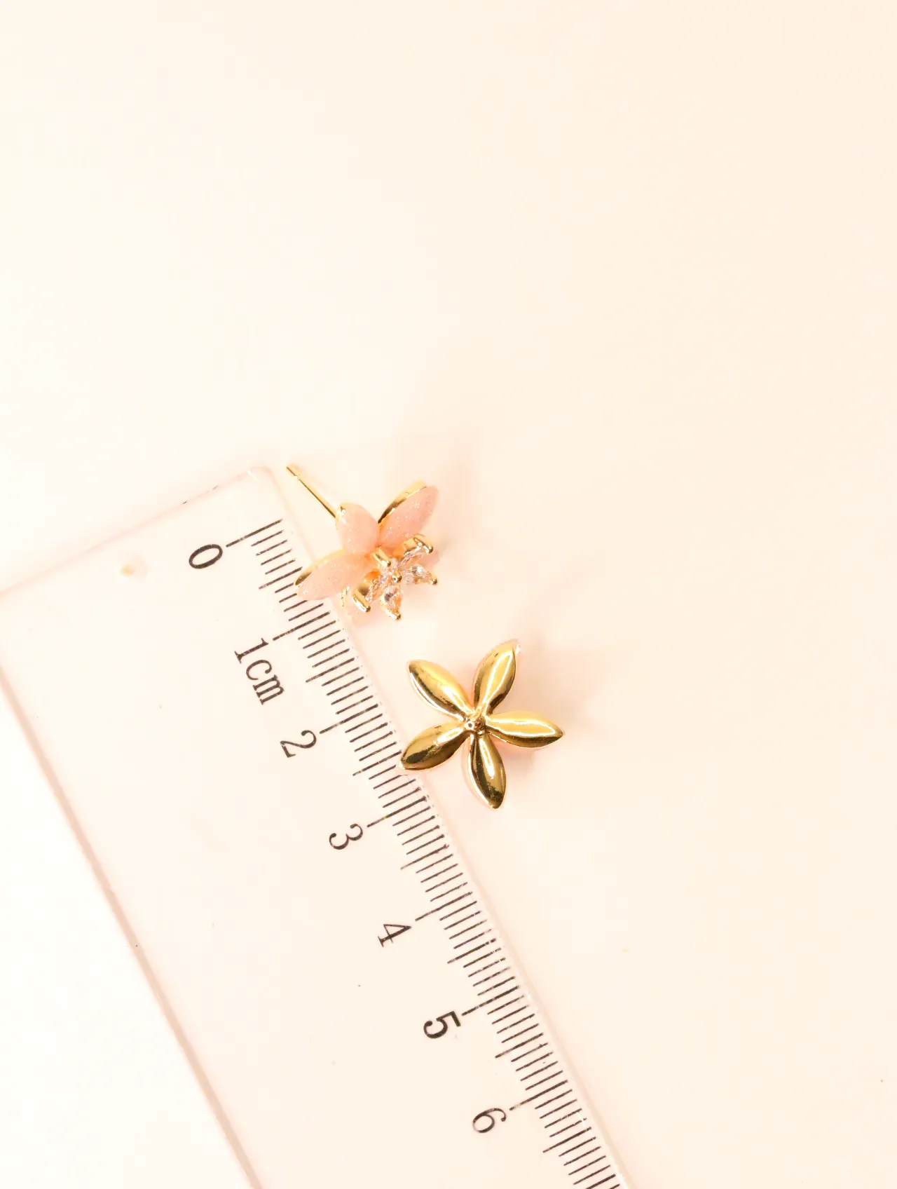 Un pendiente de botón de flor rosa junto a una regla para referencia de tamaño — pendientes dorados, primer plano · Lola y Sol (BO-PD-032)