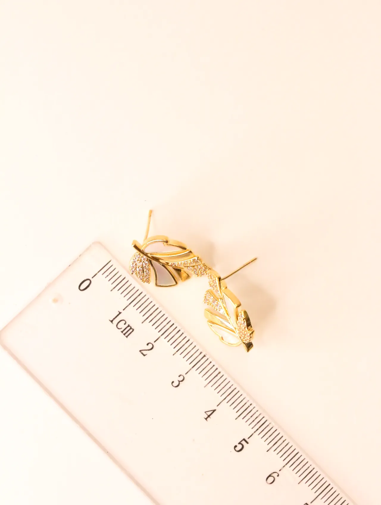Pendientes de botón dorados con diseño de hoja, nácar blanco y acentos de circonita junto a una regla, mostrando su tamaño. — pendientes dorados, primer…