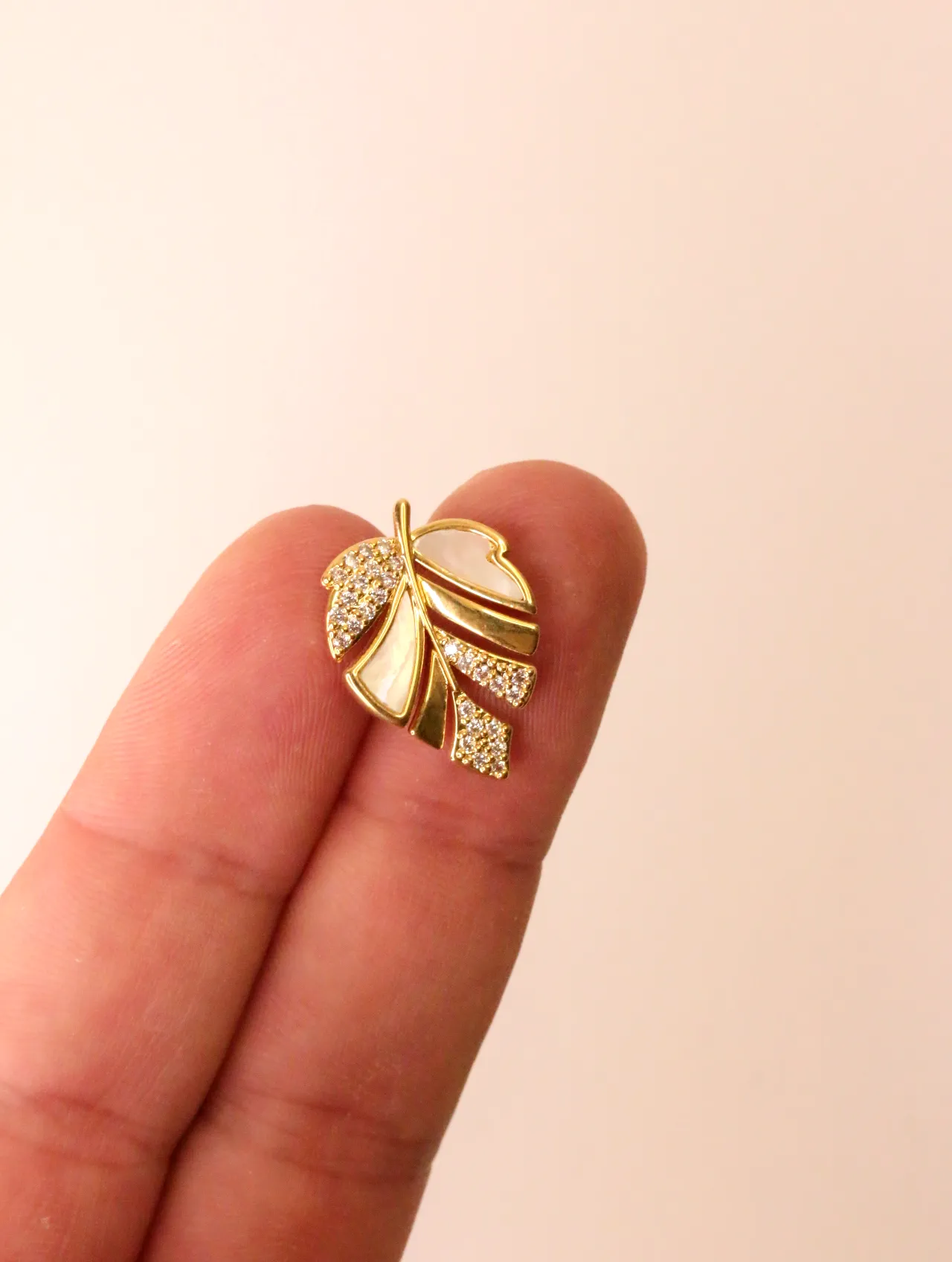 Primer plano de un único pendiente de botón dorado con diseño de hoja, nácar blanco y acentos de circonita, sostenido entre dos dedos. — pendientes orig…