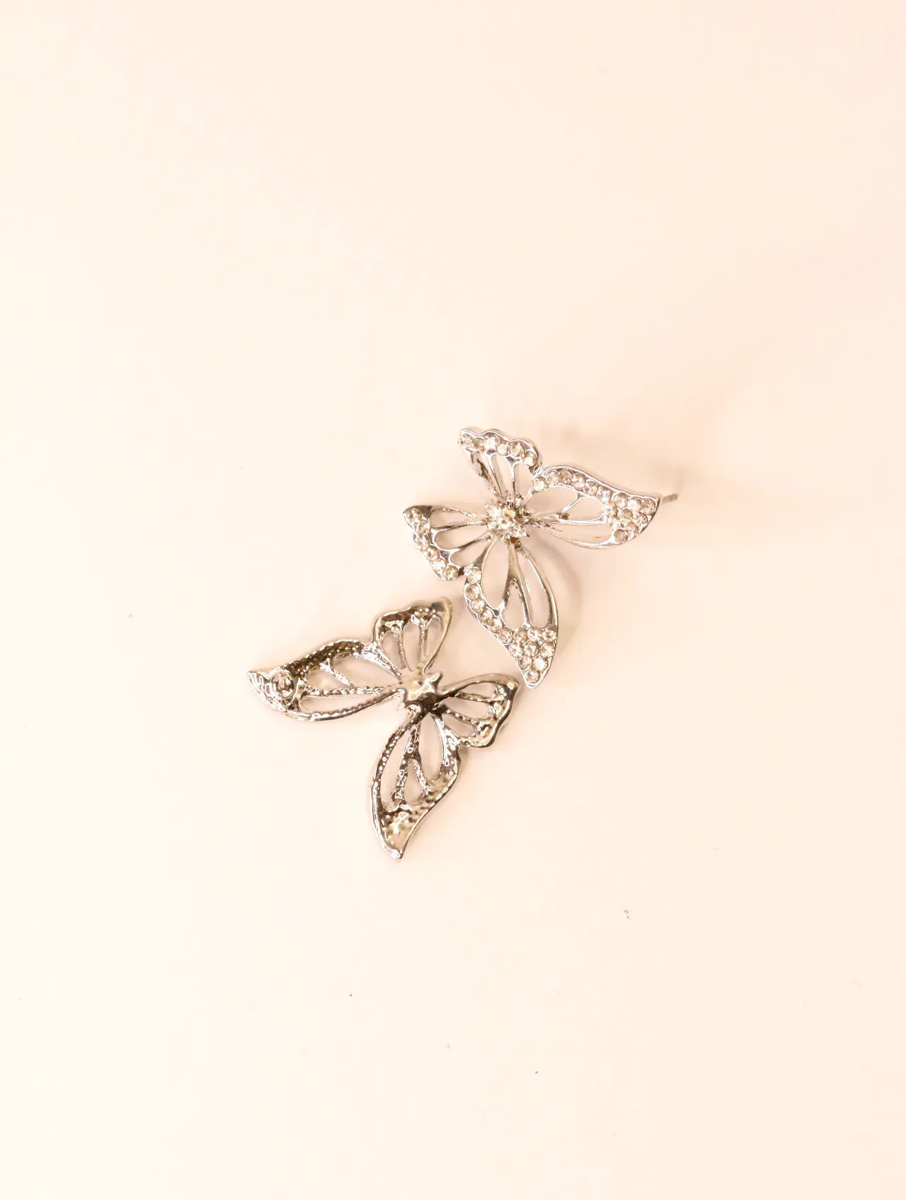 Vista superior de dos pendientes de mariposa tono plata con pedrería transparente, ligeramente superpuestos. — pendientes baratos, detalle · Lola y Sol…