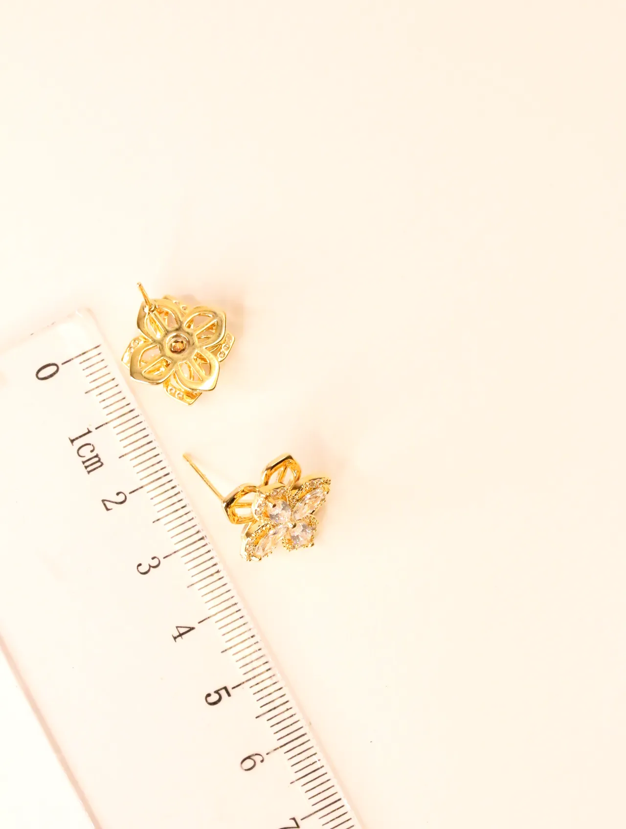 Dos pendientes de trébol tono dorado con piedras transparentes, colocados junto a una regla, mostrando su pequeño tamaño. — pendientes dorados, primer p…