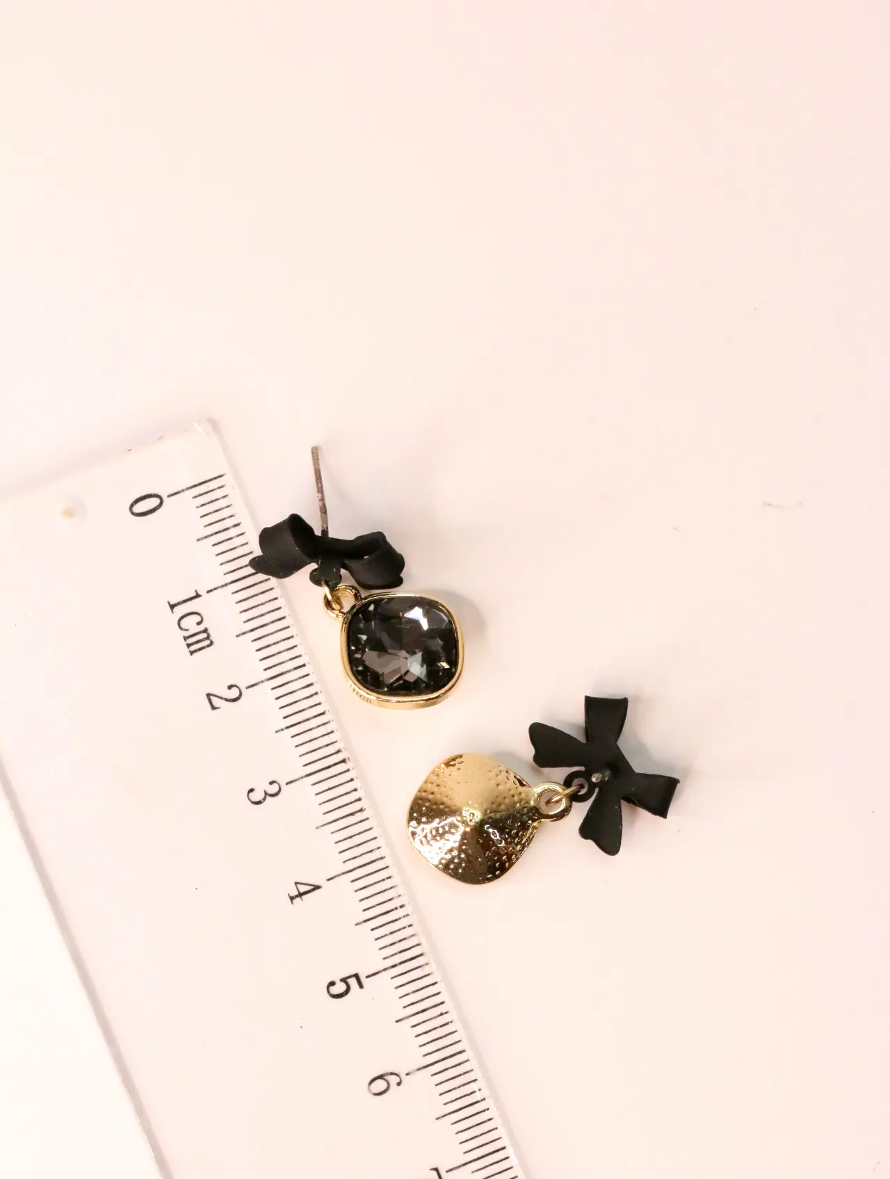 Pendientes con lazo negro y cristal junto a una regla milimetrada — pendientes dorados, primer plano · Lola y Sol (EAR-029)