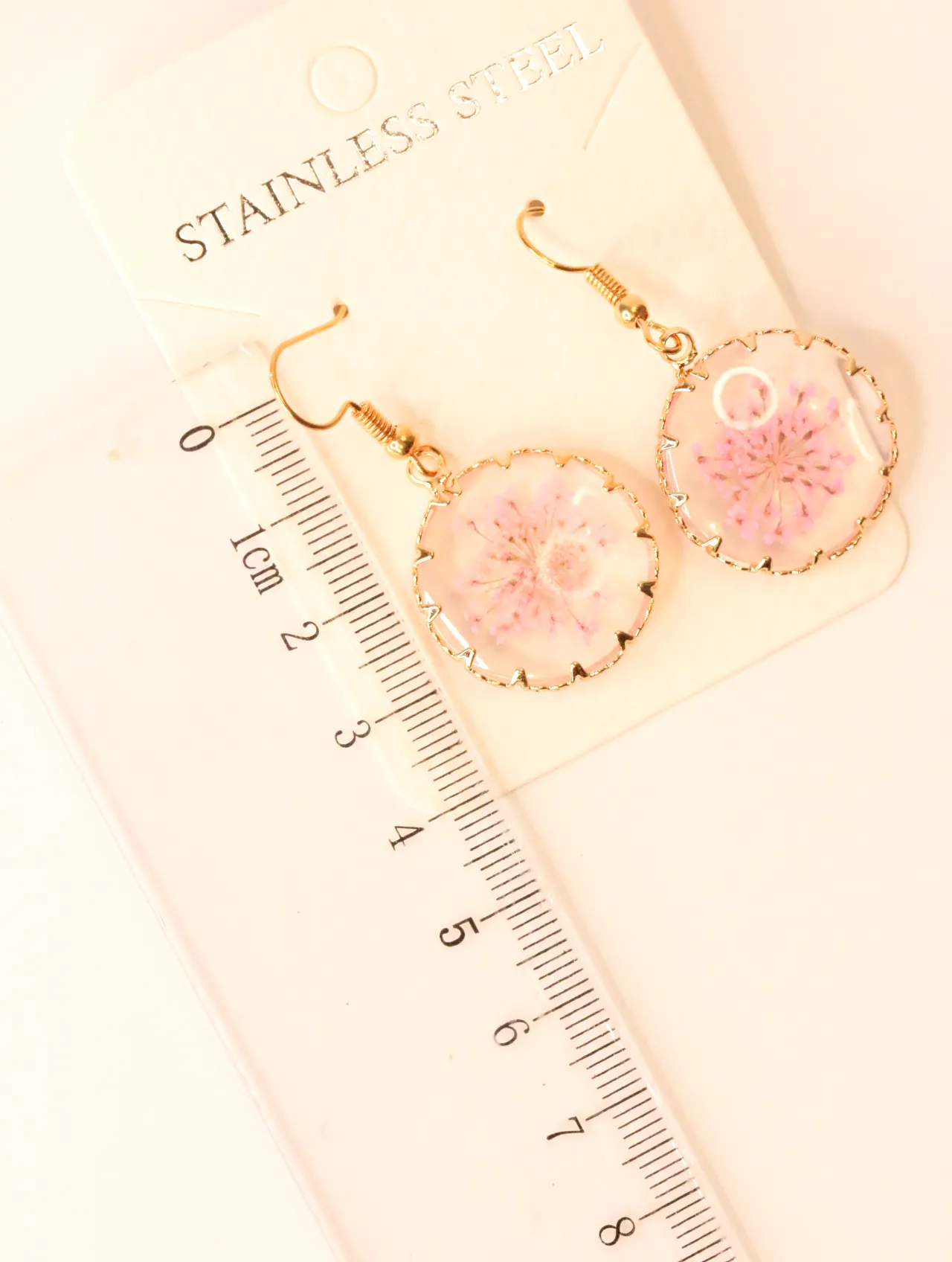 Pendientes de flores prensadas junto a una regla para referencia de tamaño — pendientes dorados, primer plano · Lola y Sol (EAR-069)