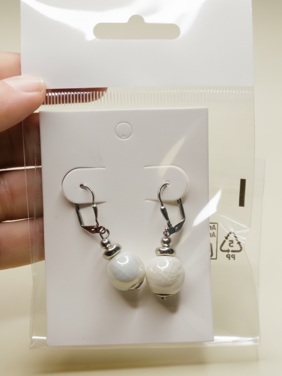 Pendientes de cuenta blanca sobre tarjeta dentro de un envoltorio transparente — pendientes dorados, primer plano · Lola y Sol (EAR-092)
